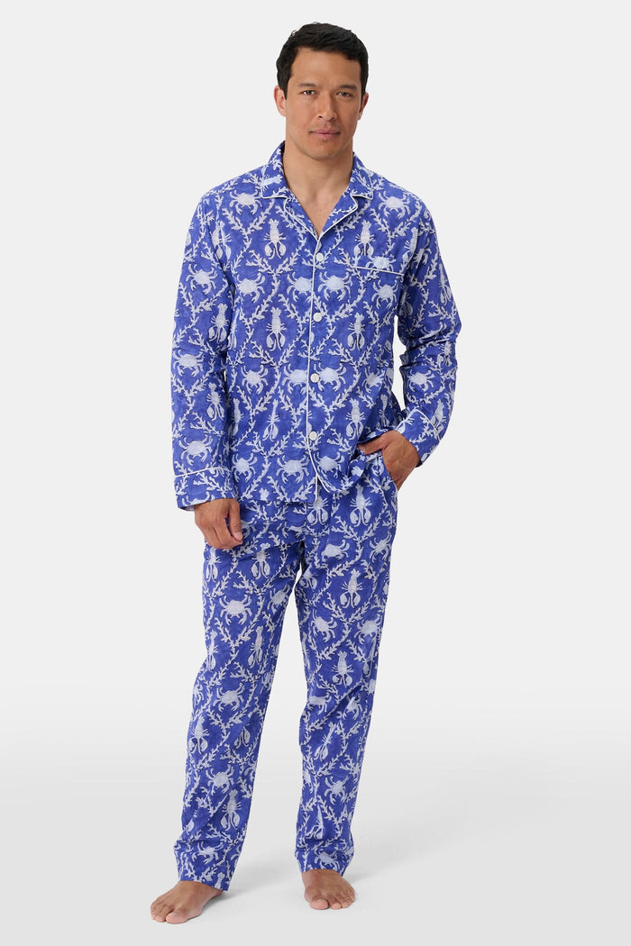 A man stands barefoot in Roller Rabbit’s Mens Lobster Trellis Loungewear, a matching blue and white floral cotton voile pajama set with a button-up long-sleeve shirt and pants. He has one hand in his pocket and looks forward.