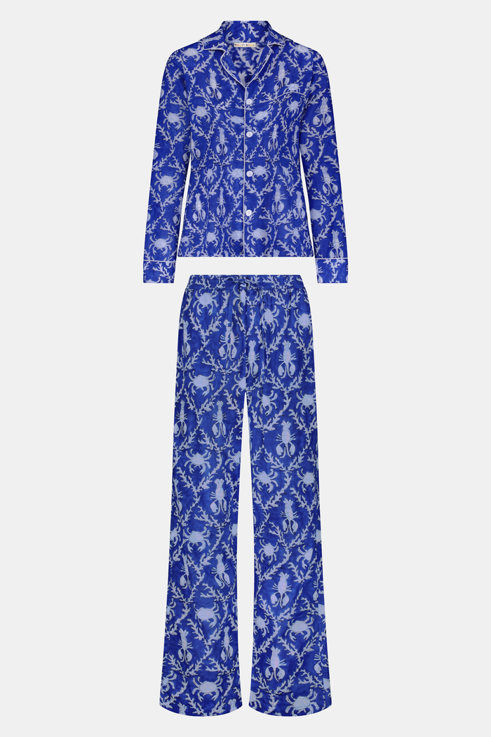 A man stands barefoot in Roller Rabbit’s Mens Lobster Trellis Loungewear, a matching blue and white floral cotton voile pajama set with a button-up long-sleeve shirt and pants. He has one hand in his pocket and looks forward.