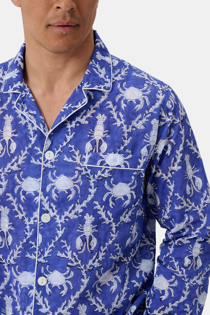 A man stands barefoot in Roller Rabbit’s Mens Lobster Trellis Loungewear, a matching blue and white floral cotton voile pajama set with a button-up long-sleeve shirt and pants. He has one hand in his pocket and looks forward.