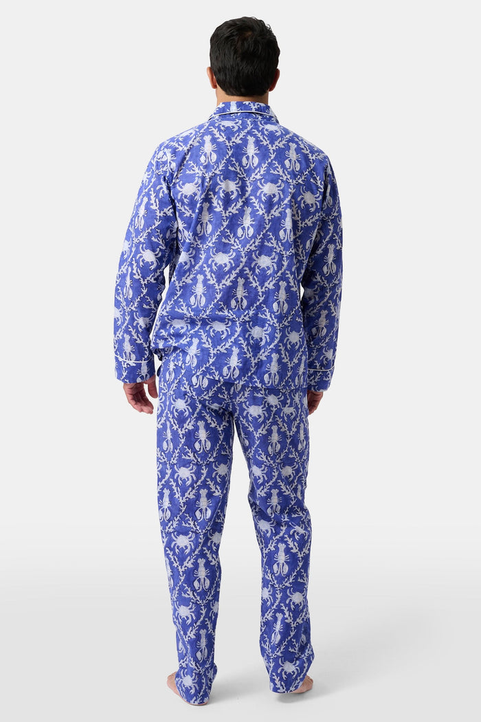 A man stands barefoot in Roller Rabbit’s Mens Lobster Trellis Loungewear, a matching blue and white floral cotton voile pajama set with a button-up long-sleeve shirt and pants. He has one hand in his pocket and looks forward.