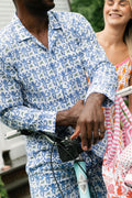 A man in Roller Rabbit's Mens Monkey Loungewear leans on a bicycle, with a smiling woman in a colorful dress standing beside him.
