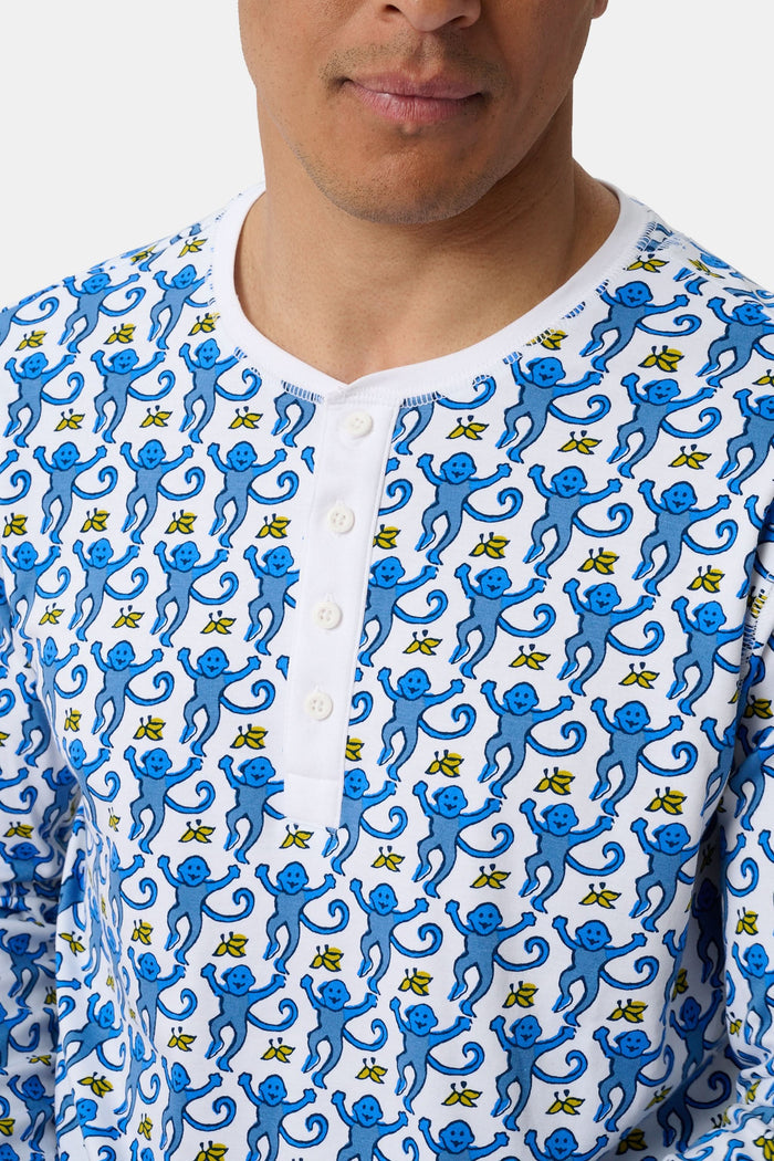 A man stands barefoot, smiling and wearing Roller Rabbit Mens Monkey Spencer Pajamas in blue and white with a playful print. The set includes a long-sleeve buttoned top and long pants. The background is plain white.