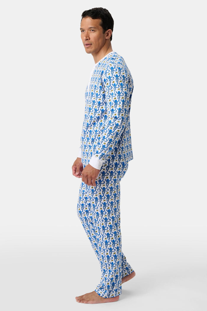 A man stands barefoot, smiling and wearing Roller Rabbit Mens Monkey Spencer Pajamas in blue and white with a playful print. The set includes a long-sleeve buttoned top and long pants. The background is plain white.