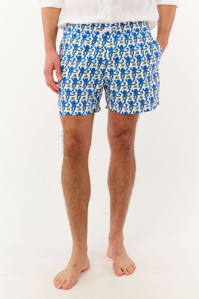 A person wearing a white shirt and Roller Rabbit’s MENS MONKEY SWIM TRUNK stands barefoot against a white background. The swim trunks showcase a playful, abstract blue-and-white design and are crafted as quick-dry swimwear with comfortable mesh-lined briefs.