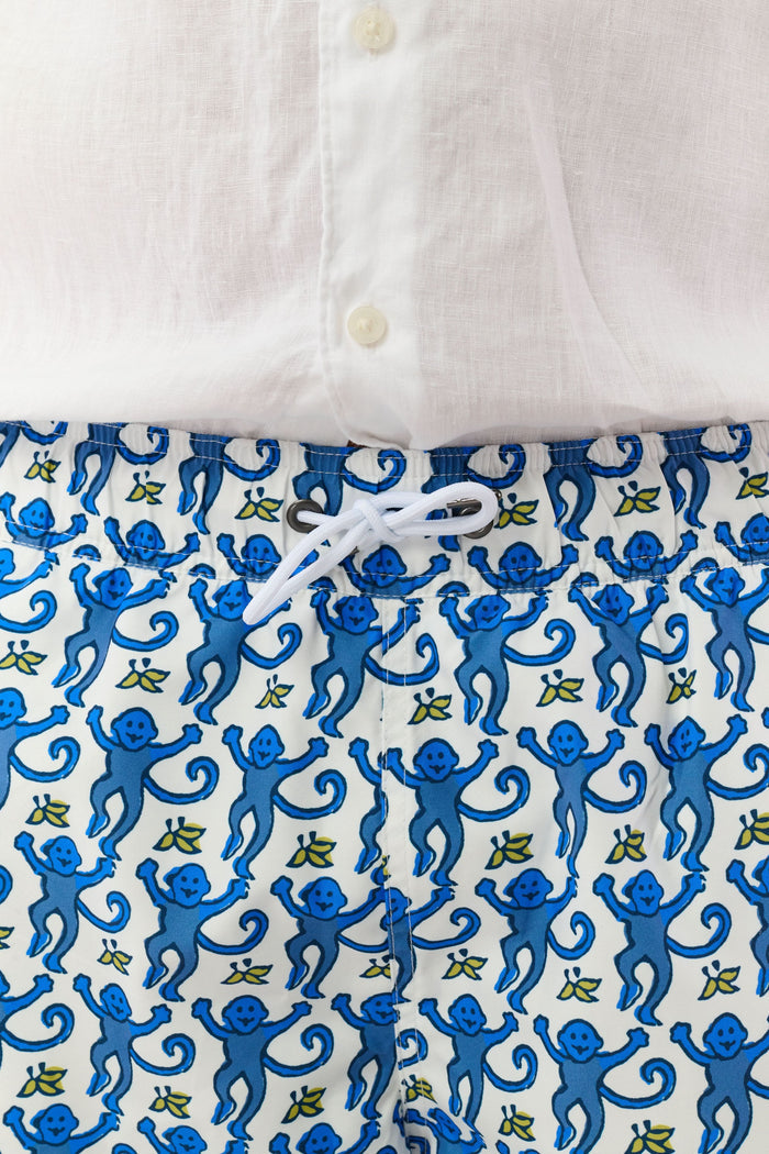 A person wearing a white shirt and Roller Rabbit’s MENS MONKEY SWIM TRUNK stands barefoot against a white background. The swim trunks showcase a playful, abstract blue-and-white design and are crafted as quick-dry swimwear with comfortable mesh-lined briefs.