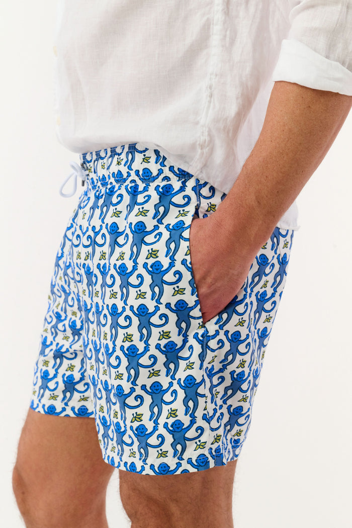 A person wearing a white shirt and Roller Rabbit’s MENS MONKEY SWIM TRUNK stands barefoot against a white background. The swim trunks showcase a playful, abstract blue-and-white design and are crafted as quick-dry swimwear with comfortable mesh-lined briefs.