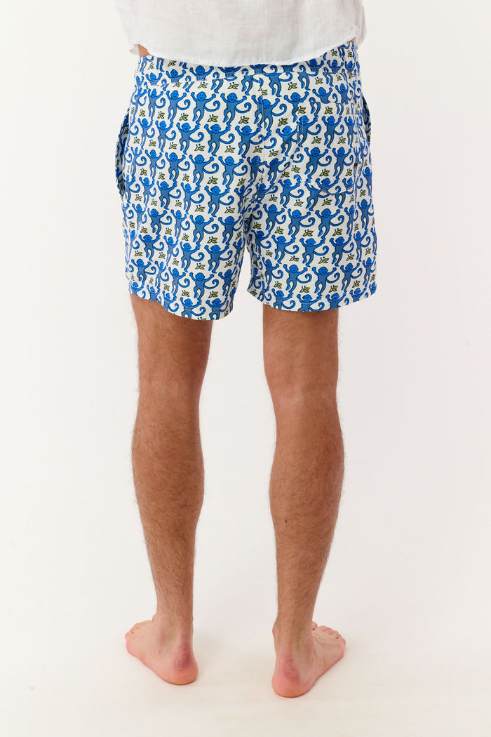 A person wearing a white shirt and Roller Rabbit’s MENS MONKEY SWIM TRUNK stands barefoot against a white background. The swim trunks showcase a playful, abstract blue-and-white design and are crafted as quick-dry swimwear with comfortable mesh-lined briefs.