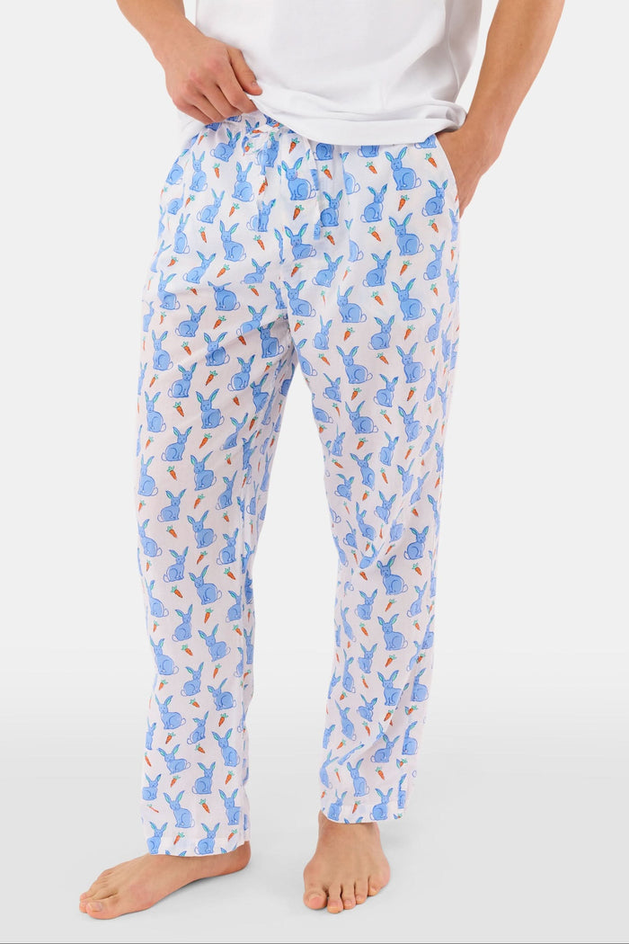 A person stands barefoot against a white background, wearing Roller Rabbit's Mens Romy the Rabbit Jon Pant—white vintage cotton pants with a blue and orange bunny print—one hand in their pocket, the other at their side.
