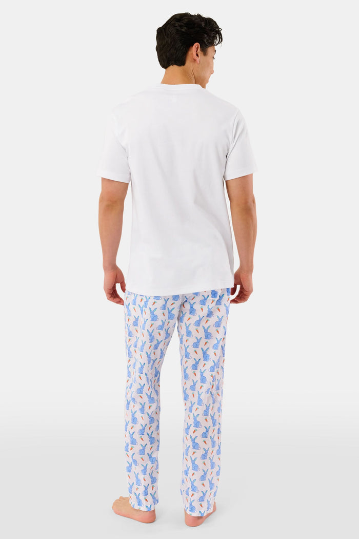A person stands barefoot against a white background, wearing Roller Rabbit's Mens Romy the Rabbit Jon Pant—white vintage cotton pants with a blue and orange bunny print—one hand in their pocket, the other at their side.