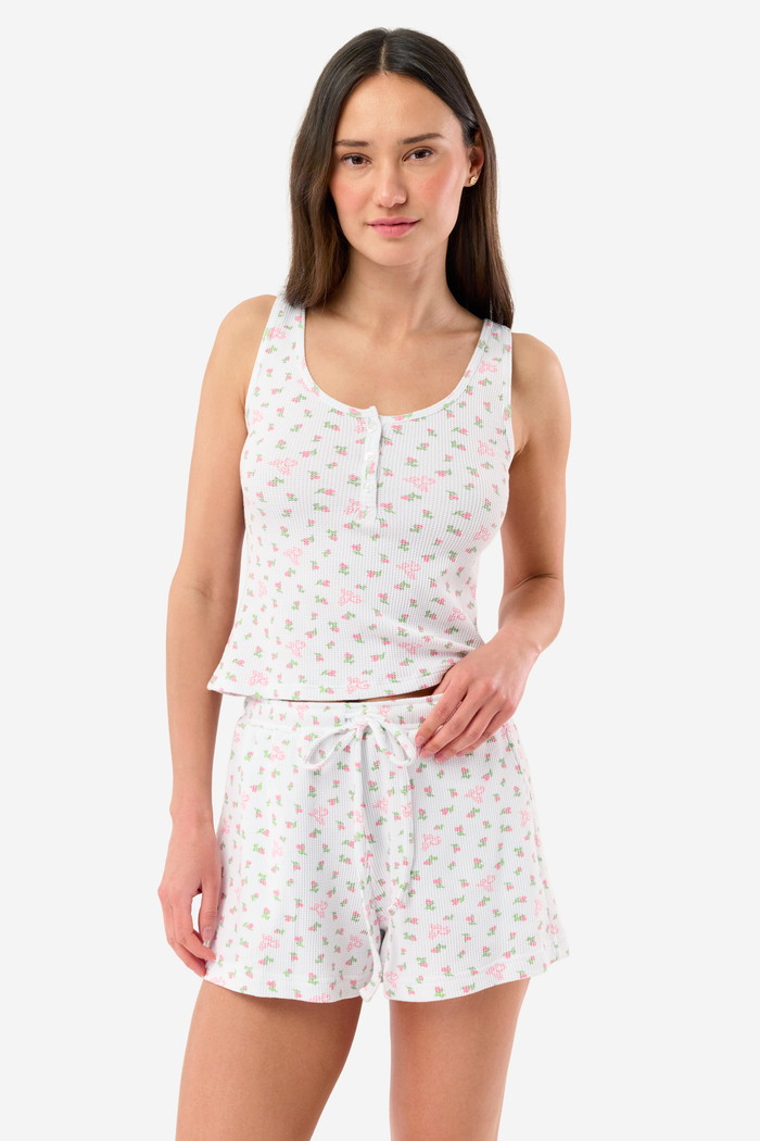 A woman stands barefoot against a white background, wearing the Roller Rabbit Mini Monkey Blossom Cherry Tank Set—a matching white tank top and shorts with a small floral pattern.