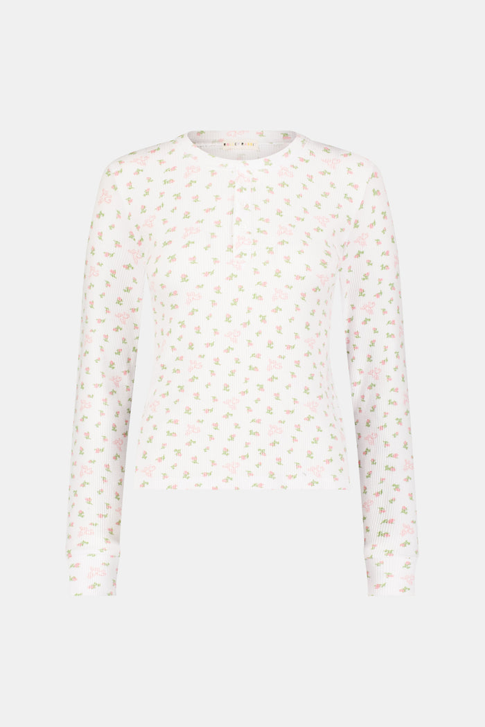 A woman wears the Mini Monkey Blossom Rosemary Top by Roller Rabbit, featuring a white pima cotton fabric with a small pink floral pattern, standing facing forward against a light background with her hands at her sides.