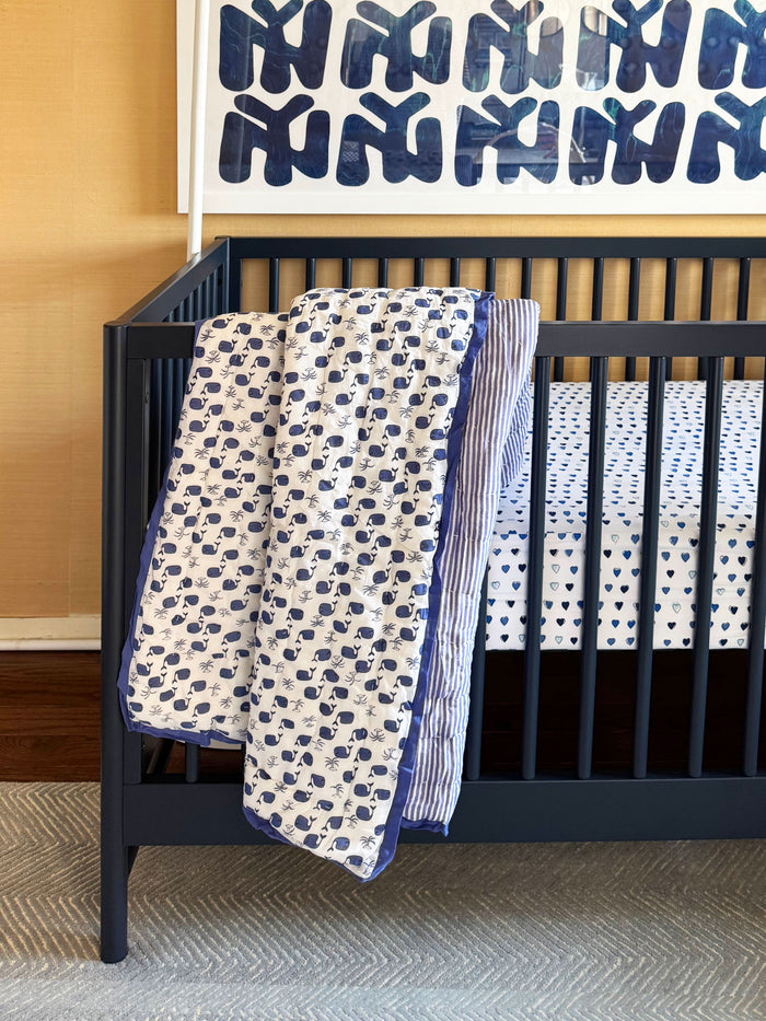 The Moby Baby Quilt by Roller Rabbit is a 100 cotton baby quilt with a white background, blue whale and seaweed patterns on one side, and blue-and-white stripes on the reverse.