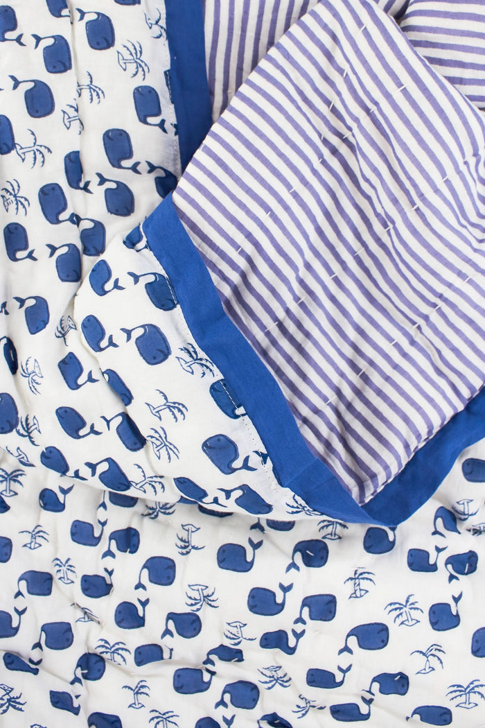 The Moby Baby Quilt by Roller Rabbit is a 100 cotton baby quilt with a white background, blue whale and seaweed patterns on one side, and blue-and-white stripes on the reverse.