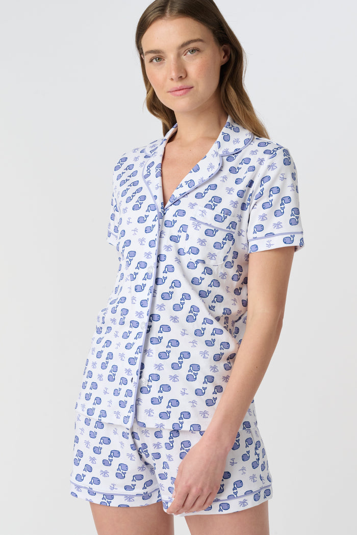 A woman with light brown hair and a gentle smile stands against a plain background, wearing Roller Rabbit's Moby Polo Pajamas—a women's short-sleeve sleepwear set featuring a whale print and crafted from soft Pima cotton.
