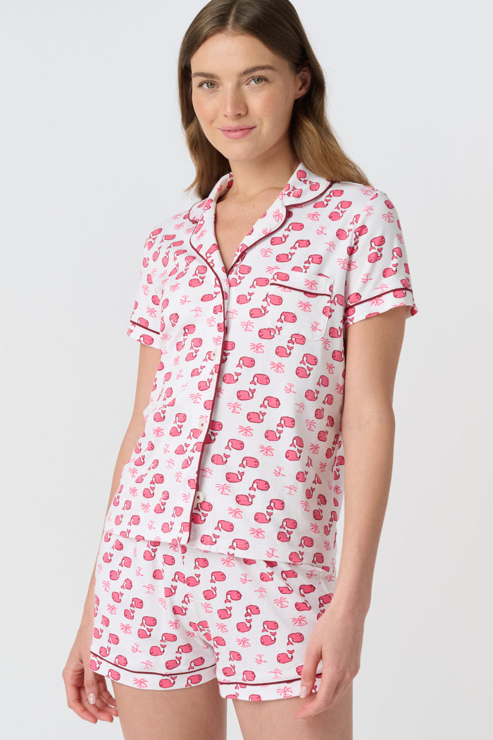 A woman wears Roller Rabbit's Moby Polo Pajamas—a women's short sleeve set with a white base and pink-red cartoon fruit prints, button-up top, and shorts. She stands against a plain light background, smiling gently.