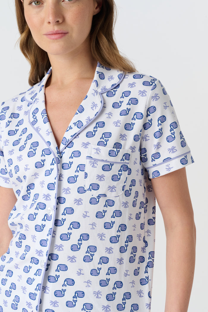 A woman with light brown hair and a gentle smile stands against a plain background, wearing Roller Rabbit's Moby Polo Pajamas—a women's short-sleeve sleepwear set featuring a whale print and crafted from soft Pima cotton.