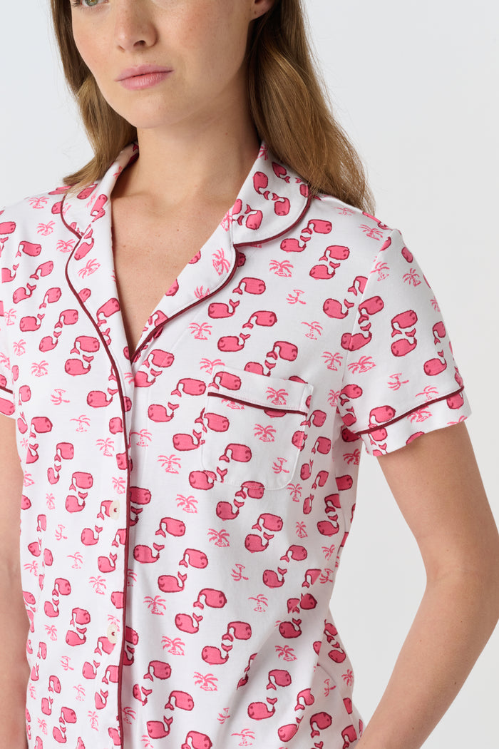 A woman wears Roller Rabbit's Moby Polo Pajamas—a women's short sleeve set with a white base and pink-red cartoon fruit prints, button-up top, and shorts. She stands against a plain light background, smiling gently.