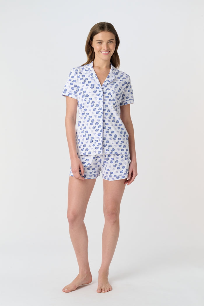 A woman with light brown hair and a gentle smile stands against a plain background, wearing Roller Rabbit's Moby Polo Pajamas—a women's short-sleeve sleepwear set featuring a whale print and crafted from soft Pima cotton.