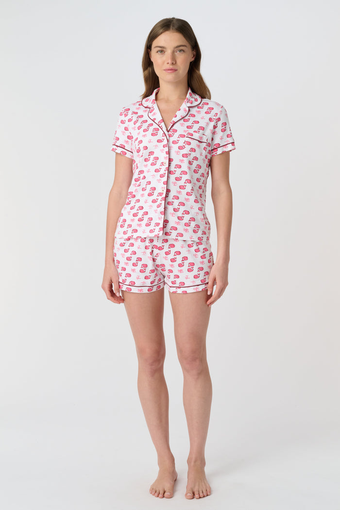 A woman wears Roller Rabbit's Moby Polo Pajamas—a women's short sleeve set with a white base and pink-red cartoon fruit prints, button-up top, and shorts. She stands against a plain light background, smiling gently.