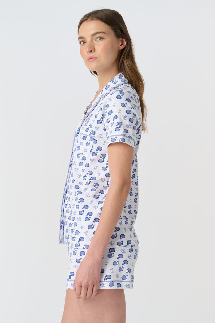 A woman with light brown hair and a gentle smile stands against a plain background, wearing Roller Rabbit's Moby Polo Pajamas—a women's short-sleeve sleepwear set featuring a whale print and crafted from soft Pima cotton.