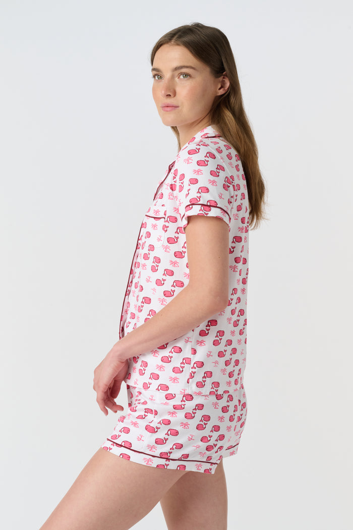 A woman wears Roller Rabbit's Moby Polo Pajamas—a women's short sleeve set with a white base and pink-red cartoon fruit prints, button-up top, and shorts. She stands against a plain light background, smiling gently.