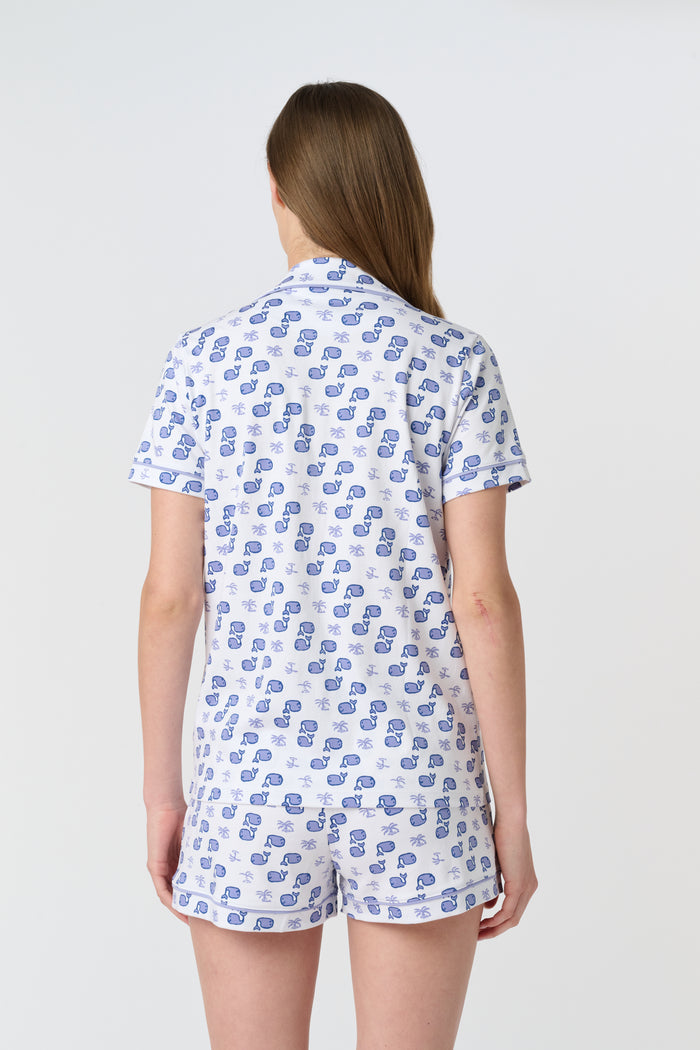 A woman with light brown hair and a gentle smile stands against a plain background, wearing Roller Rabbit's Moby Polo Pajamas—a women's short-sleeve sleepwear set featuring a whale print and crafted from soft Pima cotton.