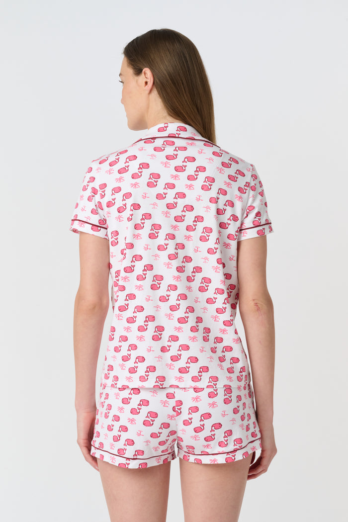 A woman wears Roller Rabbit's Moby Polo Pajamas—a women's short sleeve set with a white base and pink-red cartoon fruit prints, button-up top, and shorts. She stands against a plain light background, smiling gently.