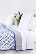 A neatly made bed features Roller Rabbit’s Moby Quilt, white pillows and sheets with blue trim, a blue and white floral blanket, and a vase of white flowers with greenery on the nightstand in the background.