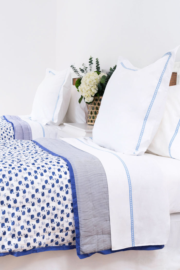 A neatly made bed features Roller Rabbit’s Moby Quilt, white pillows and sheets with blue trim, a blue and white floral blanket, and a vase of white flowers with greenery on the nightstand in the background.