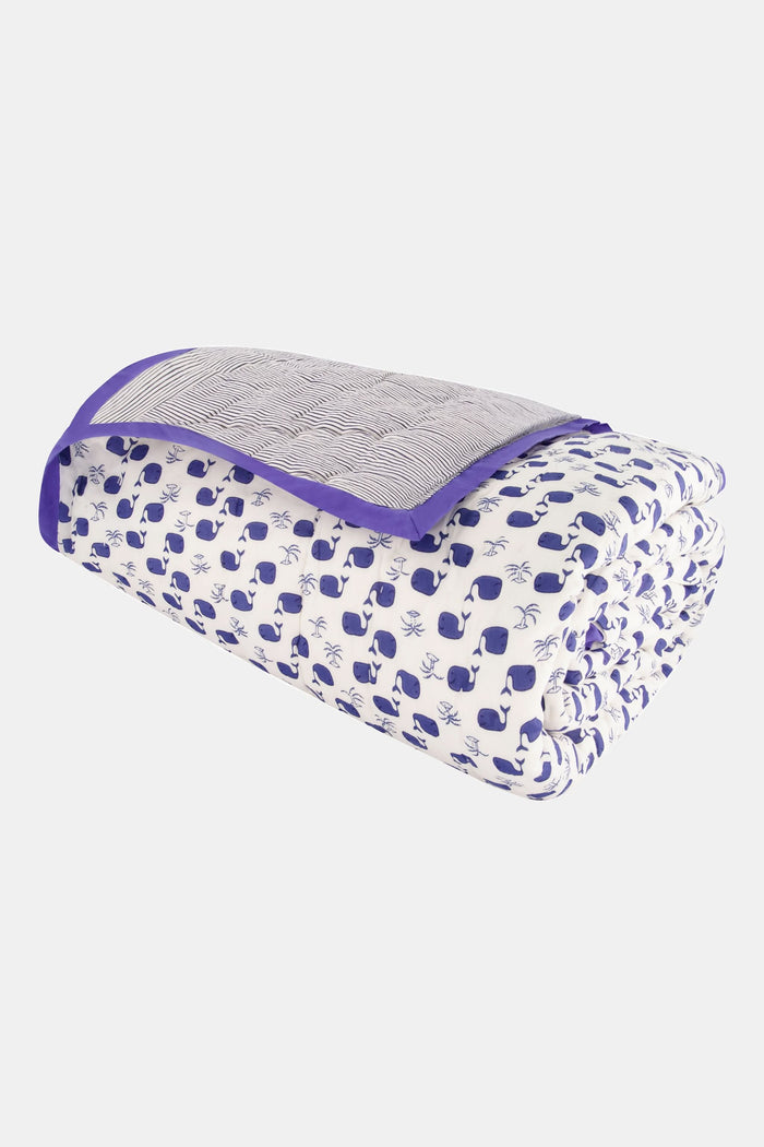 A neatly made bed features Roller Rabbit’s Moby Quilt, white pillows and sheets with blue trim, a blue and white floral blanket, and a vase of white flowers with greenery on the nightstand in the background.