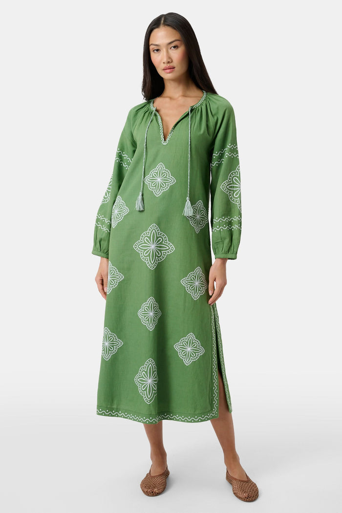 A woman wears the Roller Rabbit Monaco Geo Shiv Caftan in olive green cotton-linen with white embroidery, tassel ties at the neck, and side slits. She stands facing forward with straight dark hair and brown woven flats.