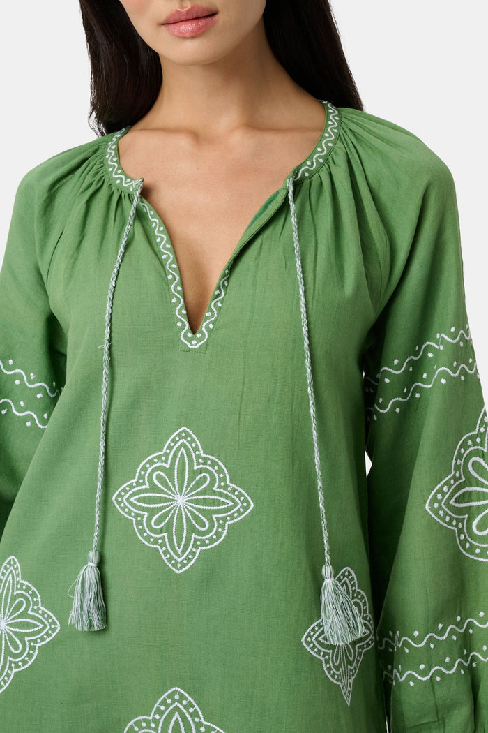 A woman wears the Roller Rabbit Monaco Geo Shiv Caftan in olive green cotton-linen with white embroidery, tassel ties at the neck, and side slits. She stands facing forward with straight dark hair and brown woven flats.