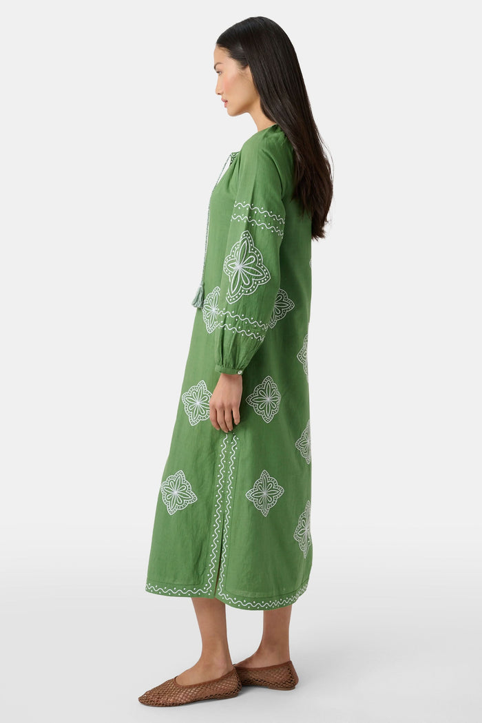 A woman wears the Roller Rabbit Monaco Geo Shiv Caftan in olive green cotton-linen with white embroidery, tassel ties at the neck, and side slits. She stands facing forward with straight dark hair and brown woven flats.