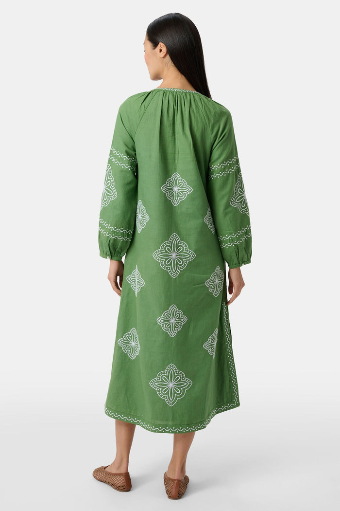 A woman wears the Roller Rabbit Monaco Geo Shiv Caftan in olive green cotton-linen with white embroidery, tassel ties at the neck, and side slits. She stands facing forward with straight dark hair and brown woven flats.