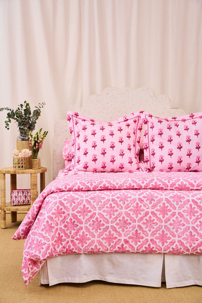 A neatly made bed features the Roller Rabbit Monaco Pool Duvet Cover in pink and white, accented with floral pillows. A wooden side table holds a vase of greenery and flowers beside a small woven basket, set against cream curtains.