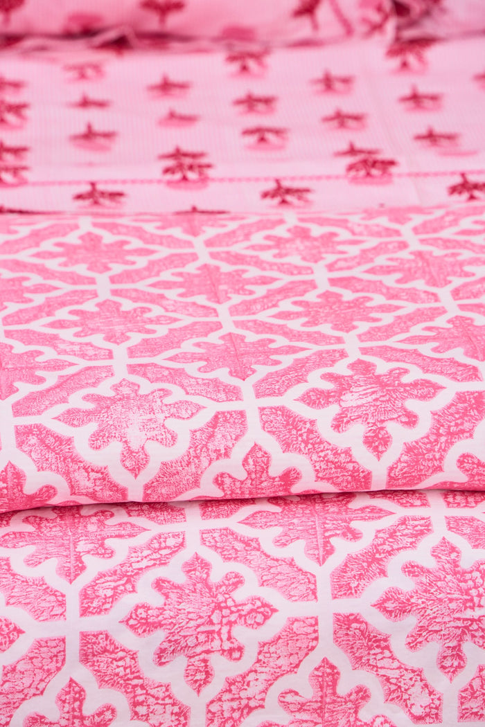 A neatly made bed features the Roller Rabbit Monaco Pool Duvet Cover in pink and white, accented with floral pillows. A wooden side table holds a vase of greenery and flowers beside a small woven basket, set against cream curtains.