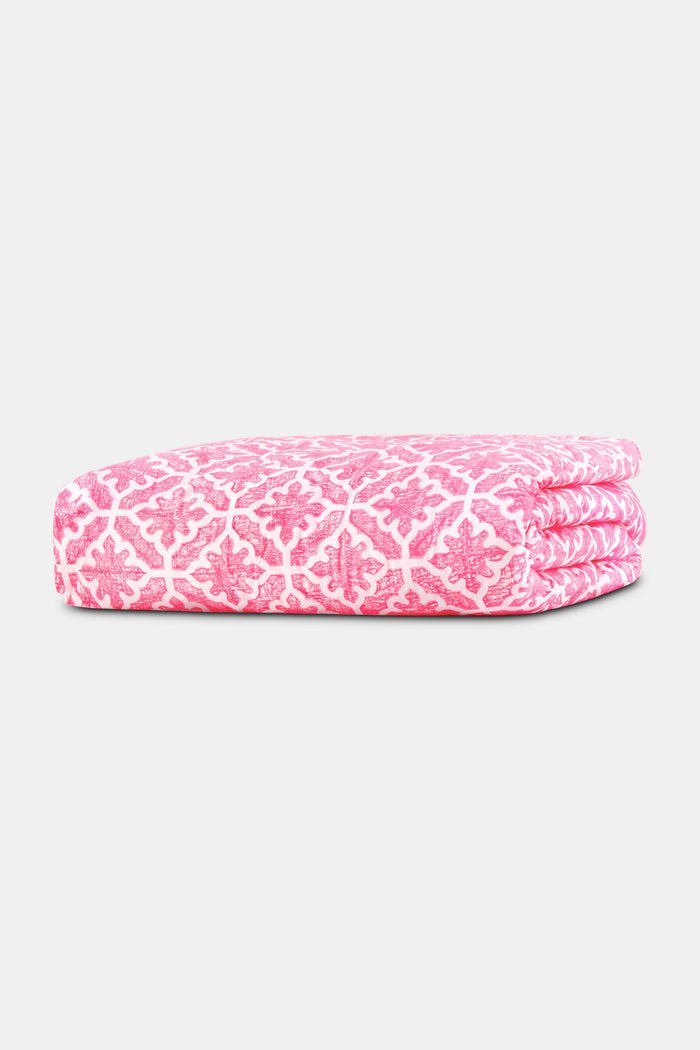A neatly made bed features the Roller Rabbit Monaco Pool Duvet Cover in pink and white, accented with floral pillows. A wooden side table holds a vase of greenery and flowers beside a small woven basket, set against cream curtains.