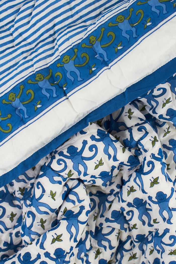 The Roller Rabbit Monkey Baby Quilt is a folded cotton quilt with a white base, blue and green monkey and leaf print, a blue border, and a decorative square featuring an intricate blue and green design.