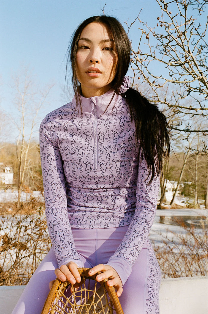 A woman with long dark hair wears the Roller Rabbit Monkey Celia Quarter Zip with thumbhole sleeves, sitting outdoors and holding a wooden snowshoe against a backdrop of bare trees and snow under a clear blue sky.