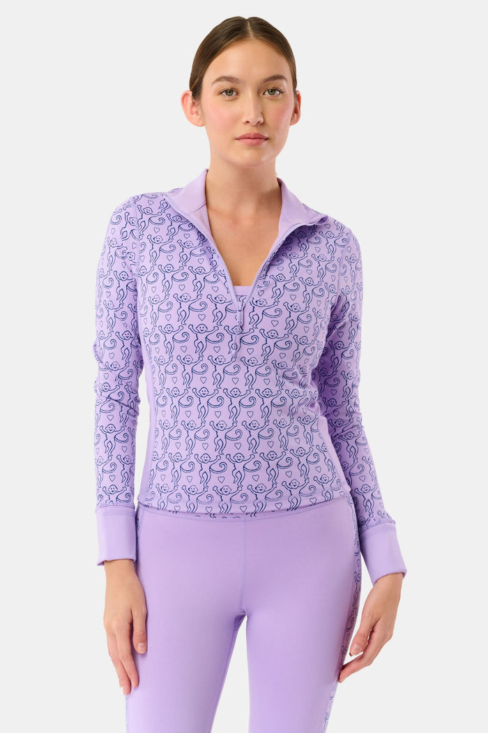A woman with long dark hair wears the Roller Rabbit Monkey Celia Quarter Zip with thumbhole sleeves, sitting outdoors and holding a wooden snowshoe against a backdrop of bare trees and snow under a clear blue sky.