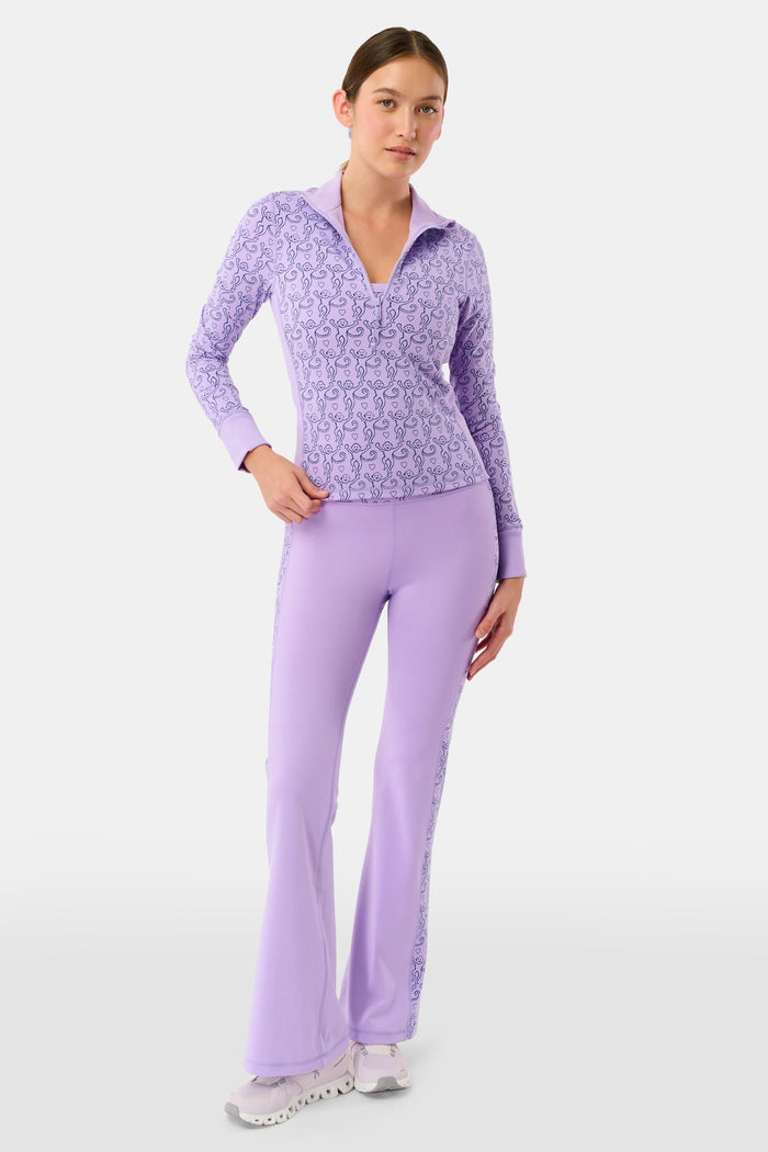 A woman with long dark hair wears the Roller Rabbit Monkey Celia Quarter Zip with thumbhole sleeves, sitting outdoors and holding a wooden snowshoe against a backdrop of bare trees and snow under a clear blue sky.