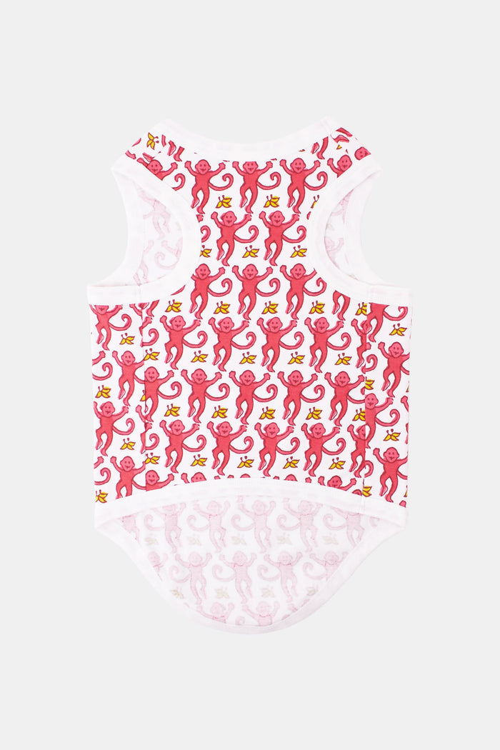 A brown poodle sits on a light surface, wearing white Roller Rabbit Monkey Dog Pajamas with a red whimsical lizard pattern, looking up and slightly to the left against a plain background.