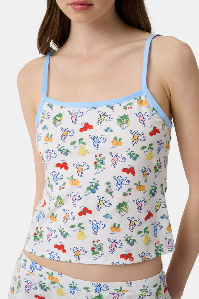 A woman wears the Monkey Fruits D’Ete Clarkson Cami by Roller Rabbit, a sleeveless white cotton-modal cami with light blue trim and a colorful fruit and cartoon print. The matching bottoms are partially visible against a plain white background.