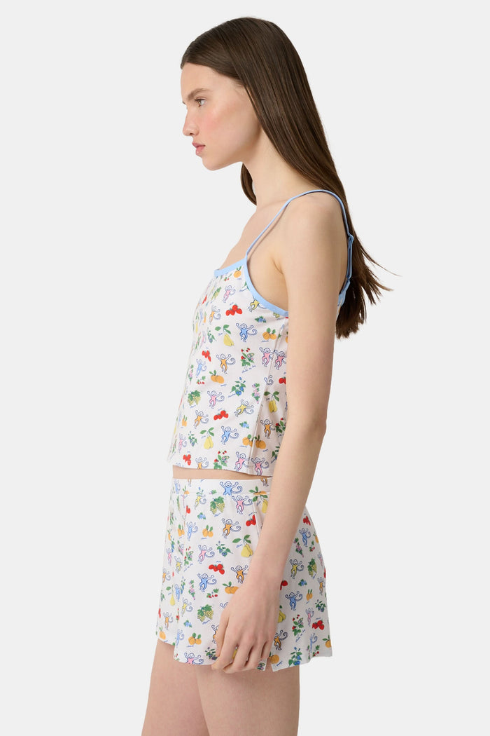 A woman wears the Monkey Fruits D’Ete Clarkson Cami by Roller Rabbit, a sleeveless white cotton-modal cami with light blue trim and a colorful fruit and cartoon print. The matching bottoms are partially visible against a plain white background.