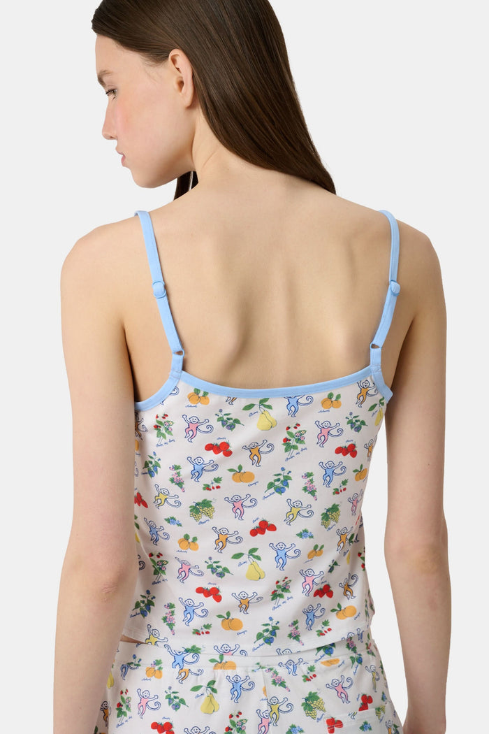 A woman wears the Monkey Fruits D’Ete Clarkson Cami by Roller Rabbit, a sleeveless white cotton-modal cami with light blue trim and a colorful fruit and cartoon print. The matching bottoms are partially visible against a plain white background.