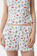 A person wearing a white sleeveless top and Roller Rabbit Monkey Fruits D'Ete Jones Shortie, featuring colorful prints of fruits, flowers, and playful cartoon figures; hands resting on the sides of the shorts.