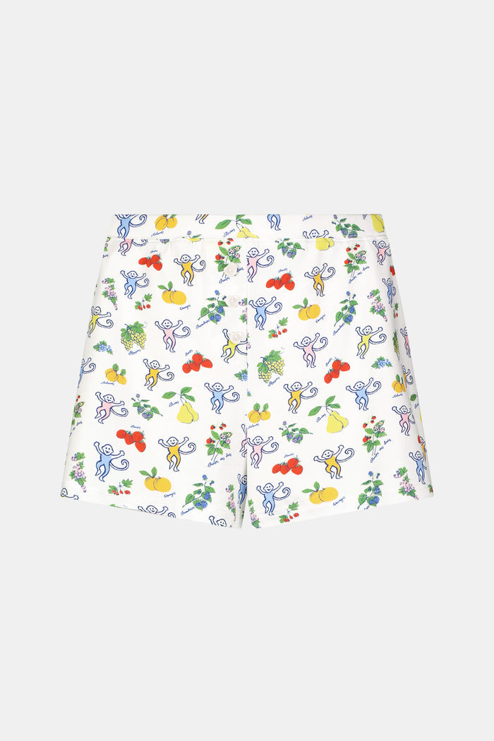 A person wearing a white sleeveless top and Roller Rabbit Monkey Fruits D'Ete Jones Shortie, featuring colorful prints of fruits, flowers, and playful cartoon figures; hands resting on the sides of the shorts.