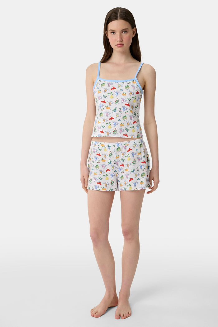 A person wearing a white sleeveless top and Roller Rabbit Monkey Fruits D'Ete Jones Shortie, featuring colorful prints of fruits, flowers, and playful cartoon figures; hands resting on the sides of the shorts.