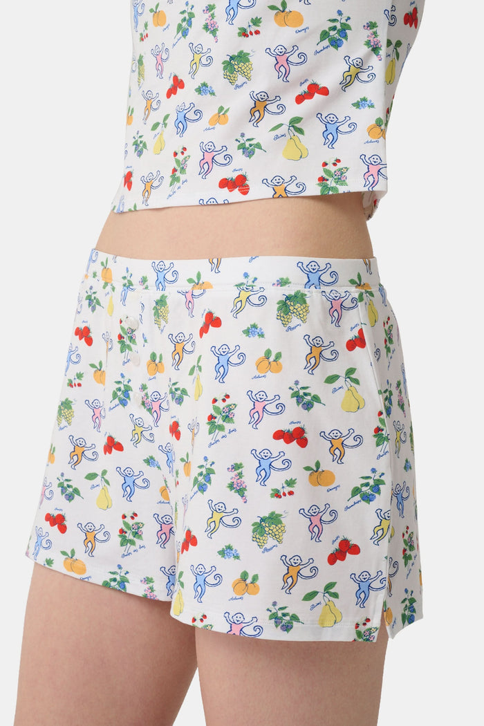 A person wearing a white sleeveless top and Roller Rabbit Monkey Fruits D'Ete Jones Shortie, featuring colorful prints of fruits, flowers, and playful cartoon figures; hands resting on the sides of the shorts.