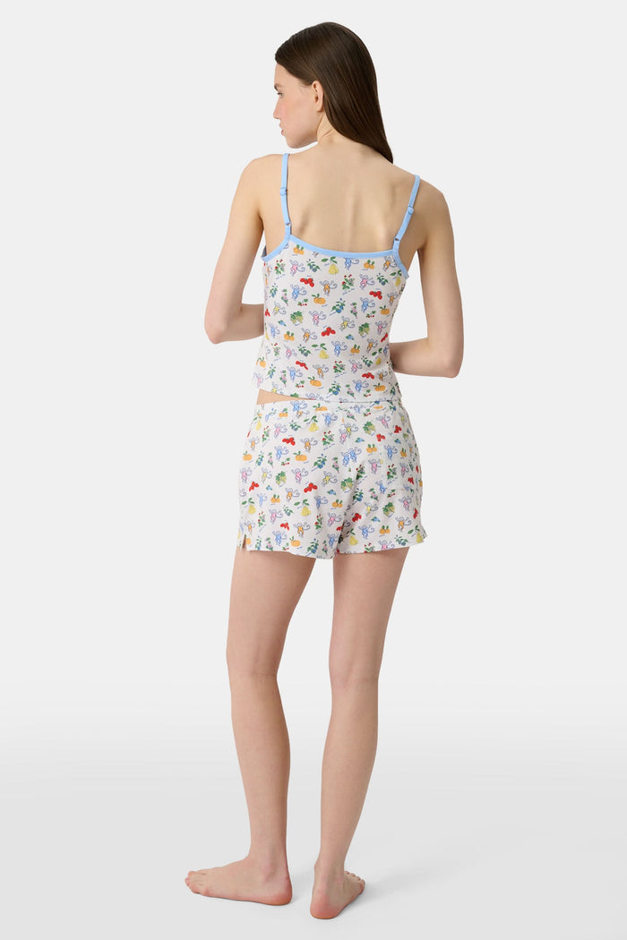 A person wearing a white sleeveless top and Roller Rabbit Monkey Fruits D'Ete Jones Shortie, featuring colorful prints of fruits, flowers, and playful cartoon figures; hands resting on the sides of the shorts.